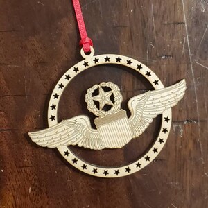 May include: A wooden ornament shaped like a circle with a star in the center and wings on either side. The ornament is decorated with a wreath and stars around the edge.