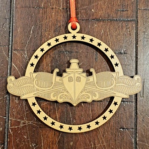 May include: A wooden ornament shaped like a submarine insignia with a red ribbon attached. The ornament is decorated with a circle of stars around the edge.