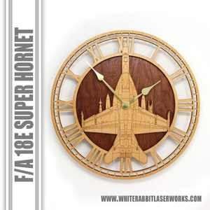 May include: A wooden wall clock with Roman numeral markings and a laser-cut design of an F/A-18E Super Hornet jet. The clock has a brown wood background and gold-colored hands.