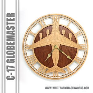 May include: Wooden wall clock featuring a C-17 Globemaster aircraft design. The clock has a light brown wooden frame with Roman numerals and a dark brown center. The clock hands are gold-colored. The text "C-17 GLOBEMASTER" is on the left side.