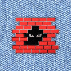 Op de afbeelding: Een rode bakstenen muur met een zwart silhouet van een gezicht met witte ogen die door een gat in de muur kijken.