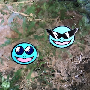 May include: Two cartoon enamel pins. One pin shows a light blue circle with large, round, blue-rimmed eyes and a smiling pink mouth. The other pin has a similar design, but with black sunglasses. Both pins have a black outline.
