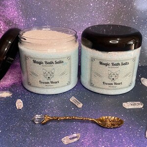 Könnte beinhalten: Zwei Glasbehälter mit Badesalz und schwarzen Deckeln. Die Behälter sind mit weißem Badesalz gefüllt und tragen ein blaues und weißes Etikett mit der Aufschrift "Magic Bath Salts by Enchantful Frozen Heart  Mistletoe".
