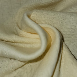 May include: Close-up of a cream-colored knitted fabric with a subtle textured pattern. The fabric is soft and drapes nicely.