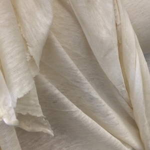 Op de afbeelding: Close-up van een witte stof met een textuurweefsel. De stof is gevouwen en gedrapeerd, waardoor de zachte en soepele textuur zichtbaar is.