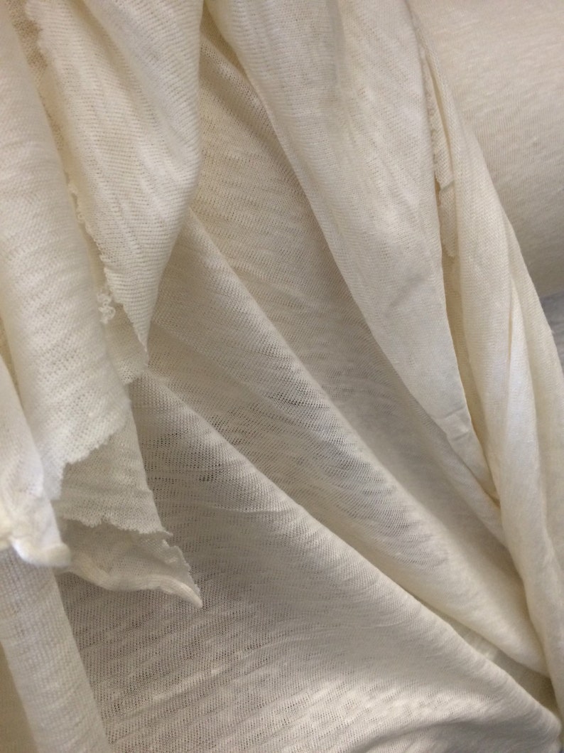 Op de afbeelding: Close-up van een witte stof met een textuurweefsel. De stof is gevouwen en gedrapeerd, waardoor de zachte en soepele textuur zichtbaar is.