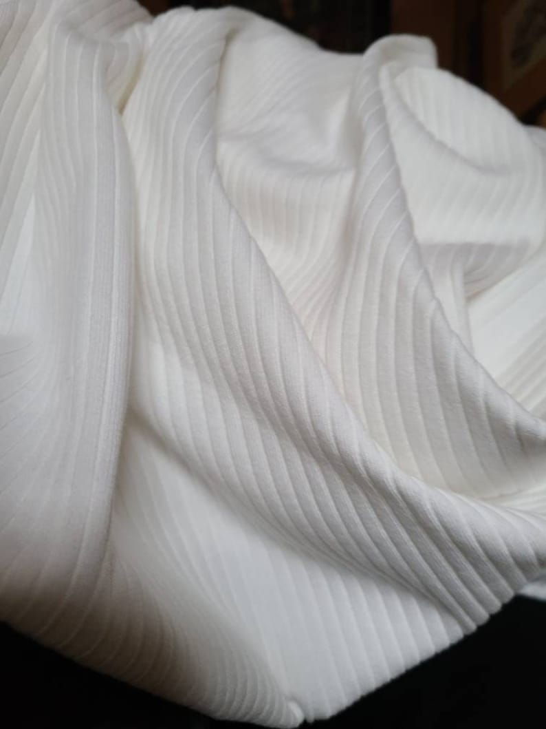 May include: White ribbed fabric with a soft, textured surface. The fabric is folded and draped, showing the texture and drape of the material.
