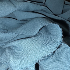 May include: Close-up of teal fabric with a textured weave. The edges of the fabric are frayed, revealing the individual threads. The fabric is folded and draped, showing the texture and color variations.