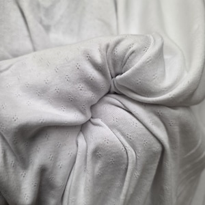 May include: A white fabric with a subtle, repeating eyelet pattern featuring small, four-pointed star-like perforations. The material is gathered and folded, creating soft, flowing drapes and shadows that highlight the texture of the textile.