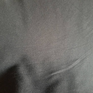 May include: Close-up of a black fabric with a ribbed texture. The fabric has horizontal lines running across its surface, creating a subtle pattern. The material appears to be soft and slightly reflective, with gentle folds and shadows.