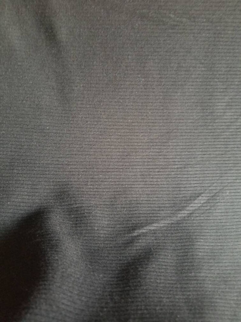 May include: Close-up of a black fabric with a ribbed texture. The fabric has horizontal lines running across its surface, creating a subtle pattern. The material appears to be soft and slightly reflective, with gentle folds and shadows.