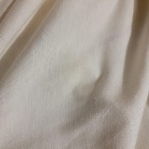 May include: Close-up of a smooth, off-white fabric with a subtle texture. The fabric has a slight sheen and is wrinkled in places.