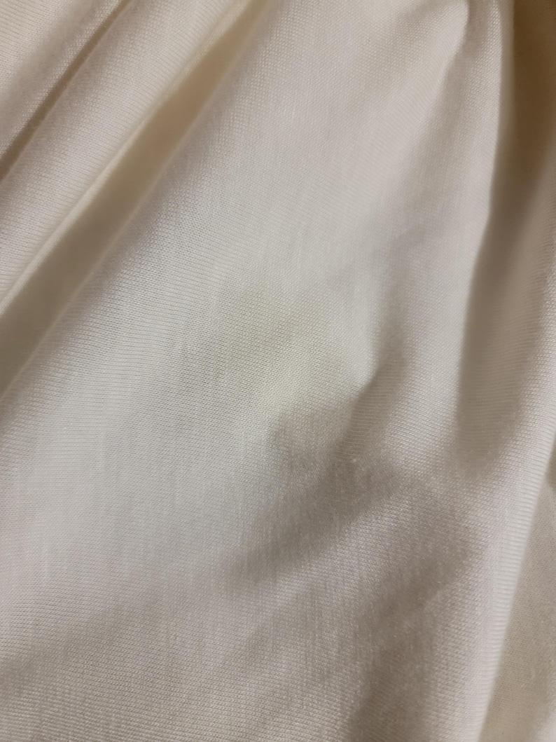 May include: Close-up of a smooth, off-white fabric with a subtle texture. The fabric has a slight sheen and is wrinkled in places.