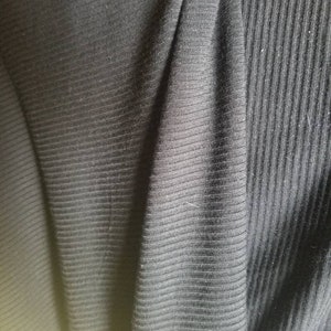 May include: Close-up of black ribbed fabric. The fabric has a textured surface with vertical lines. The material is folded, creating shadows and variations in the light.