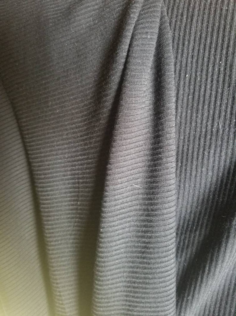 May include: Close-up of black ribbed fabric. The fabric has a textured surface with vertical lines. The material is folded, creating shadows and variations in the light.