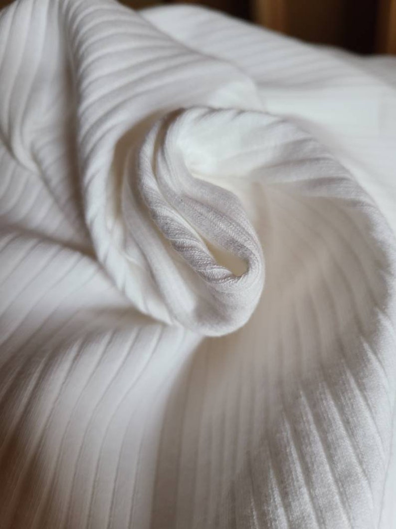 May include: White ribbed fabric with a soft, smooth texture. The fabric is folded and draped, showcasing its texture and drape.