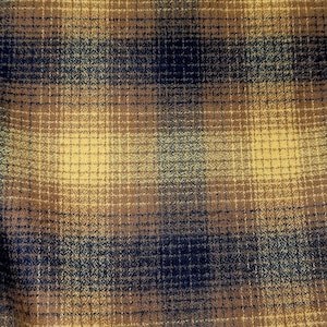 May include: Close-up of a fabric with a plaid pattern. The fabric features a blend of colors, including mustard yellow, brown, and navy blue, creating a textured appearance. The plaid design is composed of intersecting horizontal and vertical lines.