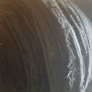 May include: A tightly rolled bolt of black fabric. The fabric appears to be a knit or woven material, with visible texture and slight fraying along the edges. The roll is tightly wound, showing the layers of the fabric.