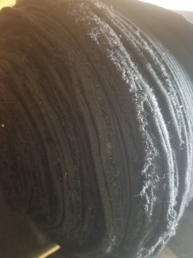 May include: A tightly rolled bolt of black fabric. The fabric appears to be a knit or woven material, with visible texture and slight fraying along the edges. The roll is tightly wound, showing the layers of the fabric.