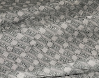 May include: Close-up of a gray and white checkered fabric. The fabric has a textured appearance with a repeating pattern of small squares and dots. The material is folded, creating shadows and depth.