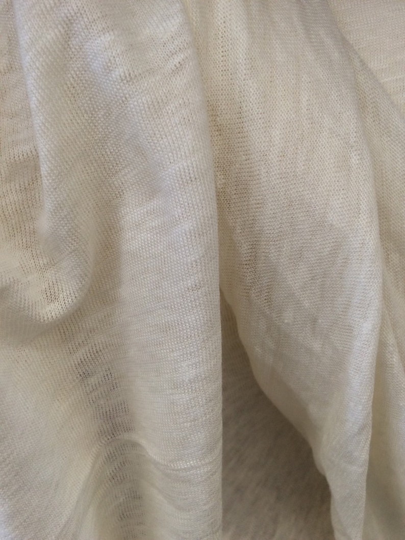 Op de afbeelding: Close-up van een witte stof met een textuurweefsel. De stof is gevouwen en gedrapeerd, waardoor de zachte, natuurlijke textuur zichtbaar is.