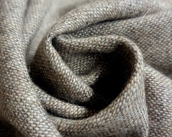 Op de afbeelding: Close-up van een gevouwen, getextureerde stof in grijstinten en bruin. Het materiaal lijkt geweven te zijn, met zichtbare draden en een zachte, licht fuzzy textuur. De plooien creëren schaduwen en diepte.
