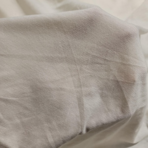 May include: Close-up of a smooth, white fabric with a subtle texture. The fabric is folded and draped, creating soft folds and curves.