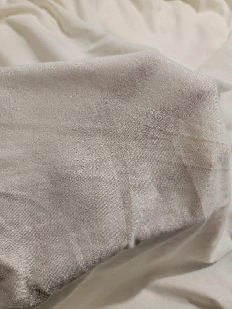 May include: Close-up of a smooth, white fabric with a subtle texture. The fabric is folded and draped, creating soft folds and curves.