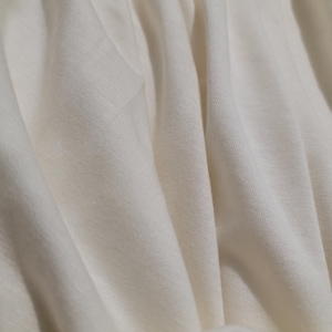 May include: Close-up of a smooth, off-white fabric with a subtle texture. The fabric is draped and folded, creating soft, flowing lines.