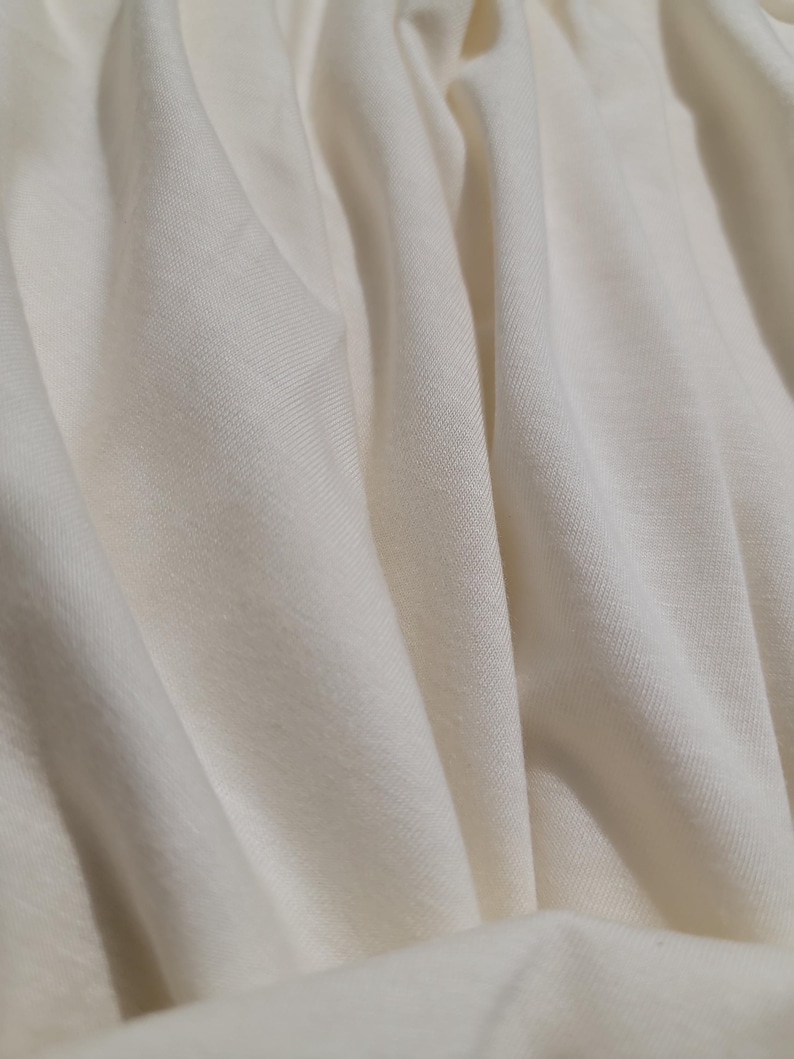 May include: Close-up of a smooth, off-white fabric with a subtle texture. The fabric is draped and folded, creating soft, flowing lines.