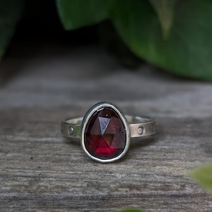 May include: A silver ring featuring a faceted, pear-shaped deep red garnet gemstone set in a smooth bezel. The metal band has a matte finish and is decorated with a series of small, evenly spaced circular indentations along the outer surface.