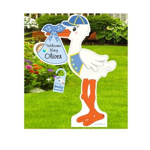 Op de afbeelding: Een blauwe en witte ooievaar die een blauwe babybundel vasthoudt met een gele strik en de tekst "Welkom Baby Oliver". Een apart blauw en wit bordje zegt "Sst... Baby slaapt!"