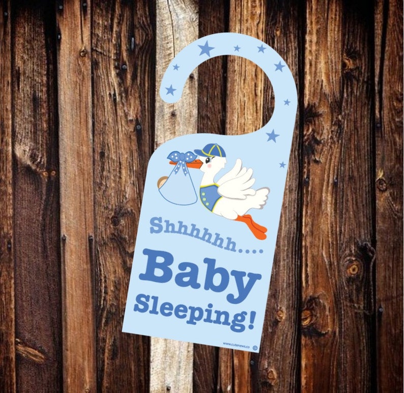 Op de afbeelding: Blauwe deurhanger met een ooievaar die een baby in een blauwe deken draagt. De hanger heeft de tekst "Shhhhh... Baby slaapt!"
