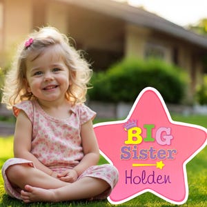May include: Pink star-shaped sign with the text "BIG Sister Holden" in colorful letters. A young girl with blonde hair is sitting in front of the sign.