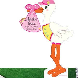 May include: A pink and white stork holding a pink and white baby bundle with a pink bow. The bundle has the text "Amelie Rose, Sept. 28, 2020, 5 lbs. 8 oz."
