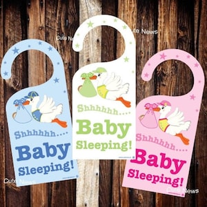 May include: Three door hangers with a stork carrying a baby. The door hangers are blue, white, and pink. The text on the door hangers reads "Shhhh... Baby Sleeping!"  Cute News.