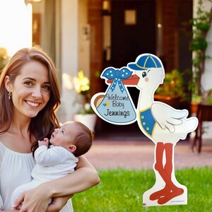 Op de afbeelding: Een witte ooievaar met een blauwe hoed en een blauwe en witte luiertas met de tekst "Welkom Baby Jennings". De ooievaar houdt de tas in zijn bek. De ooievaar staat op een groen gazon.