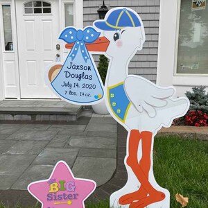 May include: A white stork holding a blue and white baby bunting with the text "Jaxson Douglas July 14, 2020 7 lbs. 14 oz."  A pink star with the text "BIG Sister" is in the foreground.
