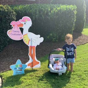 May include: A white stork holding a pink and white baby carriage with the name "Emery Catherine" written on it. A young boy wearing a blue shirt with the words "big bro" printed on it stands next to a baby car seat. A blue star with the words "Big Brother Brady" is on the ground.