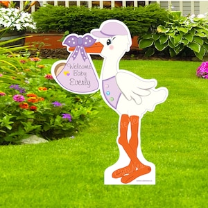 Op de afbeelding: Een paars-witte ooievaar die een paars-witte babytas vasthoudt met de tekst "Welcome Baby Everly". De ooievaar staat op een groen gazon.