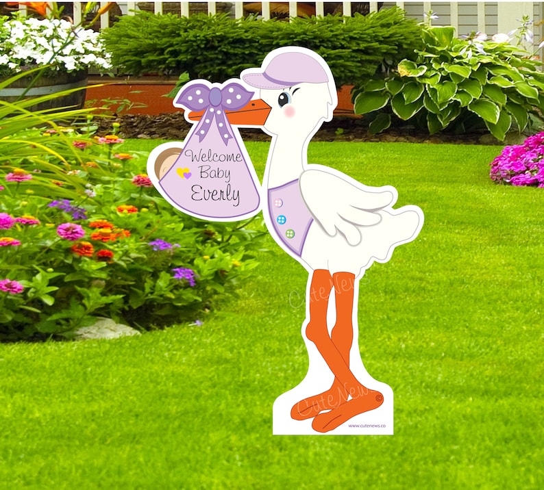 Op de afbeelding: Een paars-witte ooievaar die een paars-witte babytas vasthoudt met de tekst "Welcome Baby Everly". De ooievaar staat op een groen gazon.