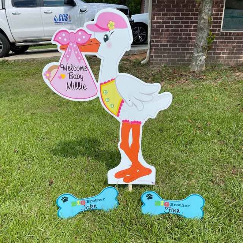 May include: A white stork holding a pink and white baby announcement sign that says "Welcome Baby Millie".  Two blue bone-shaped signs with the text "Brother Jake" and "Brother Finn" are on the ground.