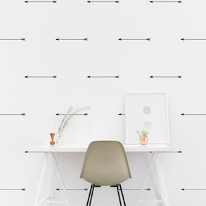 Può includere: Un muro bianco con un motivo ripetitivo di frecce nere che puntano a destra. Una scrivania bianca con una sedia bianca si trova di fronte al muro. Un quadro incorniciato è appeso al muro sopra la scrivania. Una piccola pianta si trova sulla scrivania di fronte al quadro.