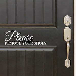 May include: A black front door with a white vinyl decal that reads "Please Remove Your Shoes". The door has a silver door handle and a silver deadbolt lock.
