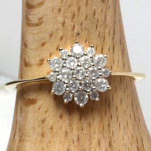 Puede incluir: Un anillo de oro con un grupo de pequeñas piedras redondas y transparentes dispuestas en un patrón circular similar a una flor. La banda es delgada y pulida, mostrando un acabado metálico brillante que contrasta con las piedras facetadas.