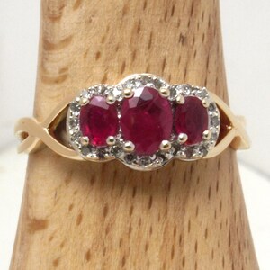 May include: A gold ring featuring three oval-cut deep red rubies arranged horizontally in the centre. The rubies are surrounded by a halo of small, clear, round-cut diamonds set in white metal, mounted on a polished gold band with a crossover design.