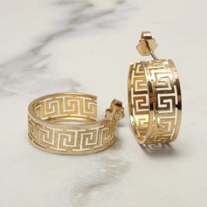 Puede incluir: Dos pendientes de aro dorados con un diseño de clave griega. Los pendientes están hechos de un metal brillante y reflectante y tienen un patrón calado. Se muestran sobre una superficie de mármol blanco.