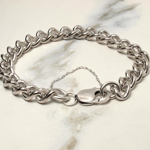 May include: A silver curb chain bracelet. The bracelet has a series of interlocking, rounded links. A lobster clasp secures the bracelet, with a small safety chain attached. The jewellery is displayed on a light-coloured surface.