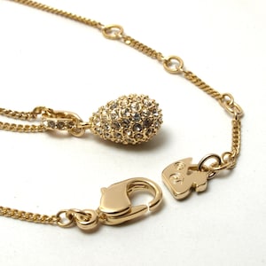 May include: A gold-tone necklace featuring a teardrop-shaped pendant encrusted with clear crystals. The chain includes a lobster claw clasp and a small swan-shaped charm near the closure, with additional small jump rings along the chain length.