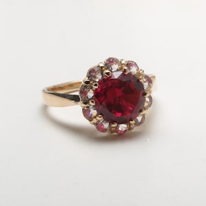 Op de afbeelding: Een gouden ring met een grote, ronde, dieprode edelsteen in het midden, omringd door kleinere, heldere en roze edelstenen. De ring heeft een eenvoudige, gepolijste gouden band.